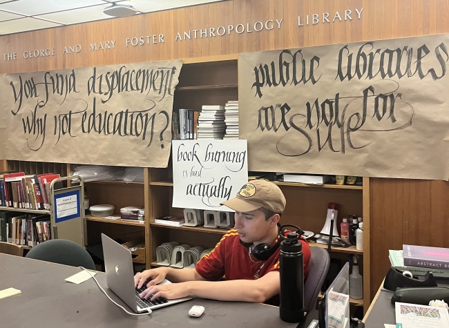 A student in a ball cap sits at a laptop with the words "The George and Mary Foster Anthropology Library" on official lettering on the wall behind him, and, on banners below those letters, “You fund displacement why not education?” and “public libraries are not for sale." 